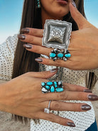 Close-up of a person wearing two ornate rings with turquoise stones. sterling silver turquoise and white buffalo adjustable cluster ring. large silver stamped concho statement ring